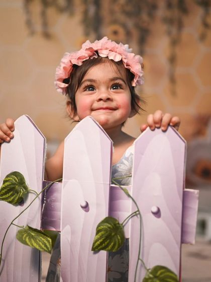 A sweet moment from a garden-themed shoot. The little girl, wearing a flower crown, peeks over a miniature picket fence, creating an adorable and interactive portrait.