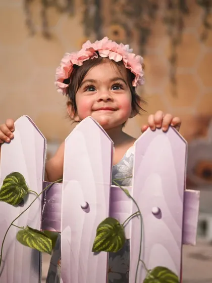 A beautiful portrait from a sunflower-themed shoot. The baby peeks over a small picket fence prop, with a soft, warm background creating a dreamy atmosphere.