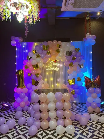 A butterfly-themed first birthday party. This setup features a balloon arch in pastel purple and pink, with butterfly cutouts and a 'Happy Birthday' neon sign.