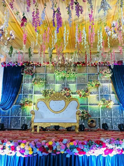 A dreamy stage setup with a ceiling of hanging wisteria, a sparkling backdrop, and royal blue drapes. The multi-colored floral border adds a pop of vibrancy.