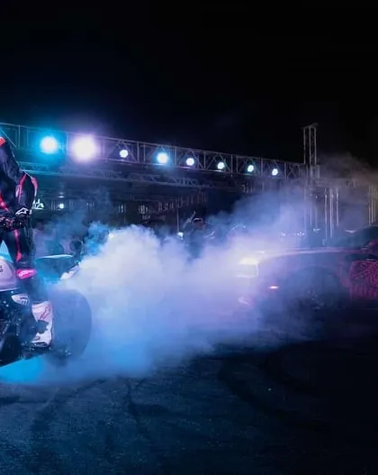We create a spectacle of light, sound, and skill. A rider performs a burnout on a custom motorcycle, with vibrant blue smoke filling the arena under the glow of professional stage lighting, captivating the crowd.