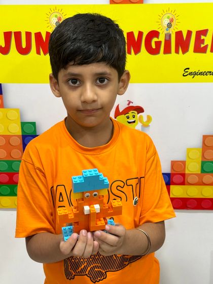 A student in an orange shirt shows off his small, creatively designed Ganesha idol made with blue and orange bricks.
