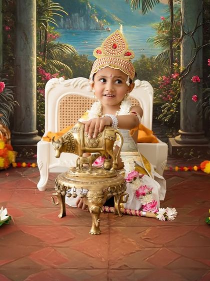 A little girl dressed as Krishna, seated on a miniature throne. The vibrant backdrop and traditional props like the brass cow and lotus flowers complete this divine setup.