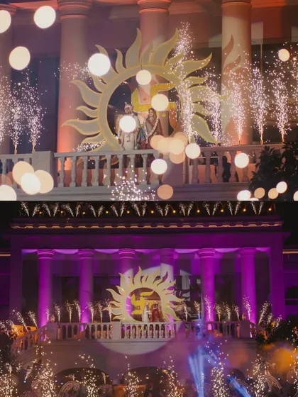 A wide shot of a spectacular varmala ceremony, with the couple framed by a giant sun prop and surrounded by cold-spark pyrotechnics.