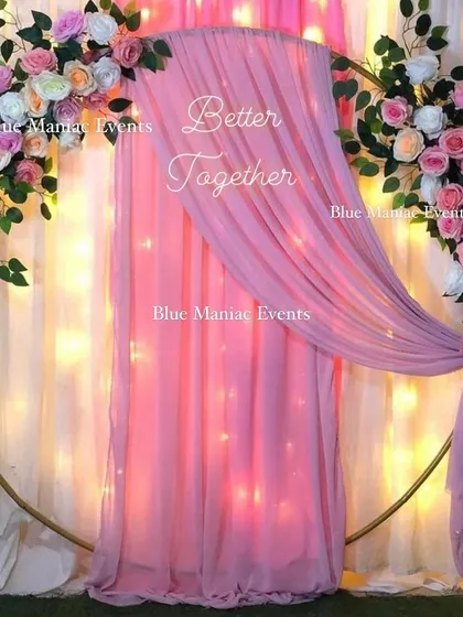 A simple yet beautiful 'Better Together' backdrop with a circular frame and pastel pink florals and draping. The fairy lights in the background add a soft, romantic glow.