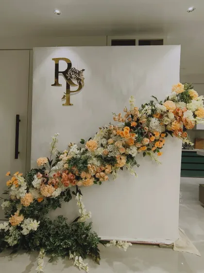 An elegant and minimalist engagement backdrop. A simple white panel is decorated with a stunning diagonal arrangement of peach and white flowers and a gold acrylic monogram of the couple's initials.