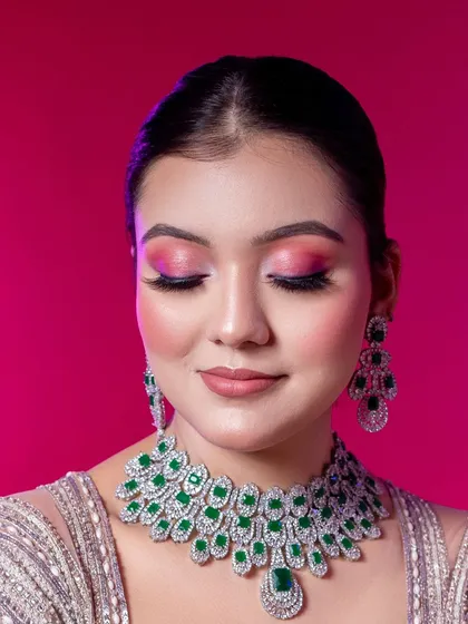 A detailed close-up shot designed to highlight technical makeup skill. The model's eyes are closed to fully display the vibrant pink and magenta eyeshadow blend. This image also showcases the stunning emerald and diamond necklace set provided as part of our styling service.