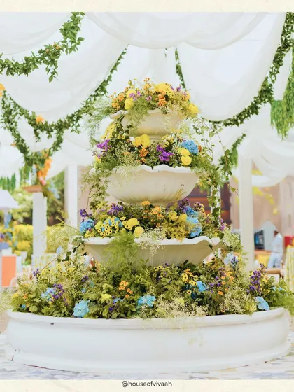 A three-tiered fountain overflowing with a vibrant mix of yellow, blue, and purple flowers, acting as a stunning central decor piece for the Mehendi.