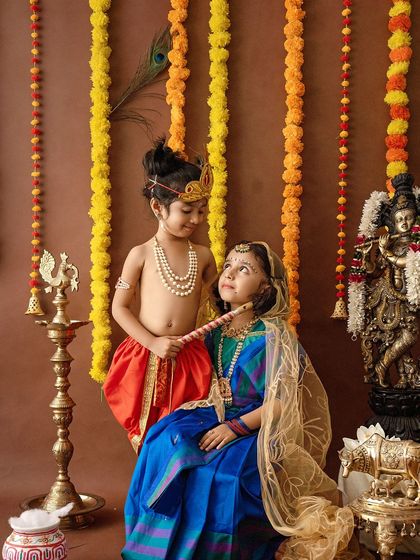 A sweet interaction between siblings dressed as Krishna and Radha. The setup includes a traditional swing, marigold garlands, and brass decor to create an authentic festive atmosphere.