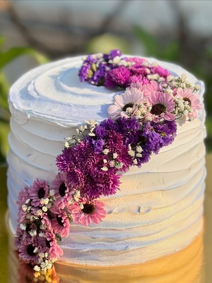 A tall cake with textured white frosting, decorated with a spiral of fresh purple and pink flowers. The color gradient of the flowers adds a beautiful visual element.
