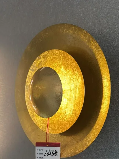 A gold-leaf wall sconce that doubles as wall art. The light emanates from behind the inner circle, creating a warm, glowing halo.