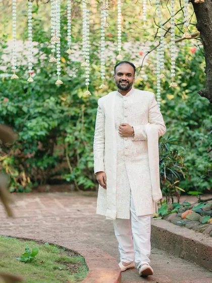 A full-length portrait of the groom in a classic white sherwani. The natural garden setting provides a perfect backdrop for this elegant and timeless shot.