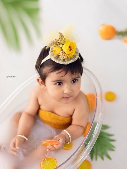 A close-up of the baby girl in her fruit bath, looking thoughtful. The details, like her tiny fingers holding an orange slice, are precious.