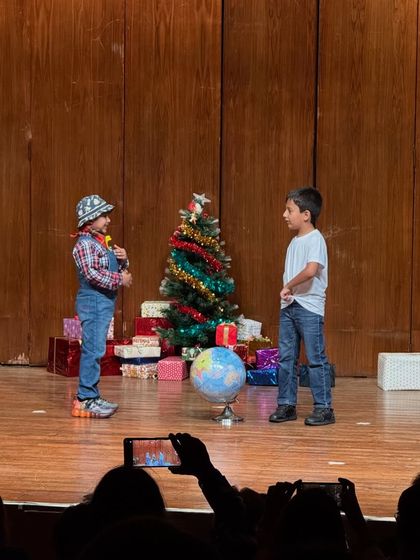A farmer character interacts with another student, with proud parents capturing the moment from the audience.