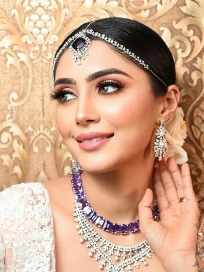 A stunning bridal look featuring a sleek, clean bun adorned with a single white flower. The hair is pulled back to showcase the intricate matha patti and dramatic makeup.