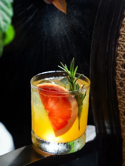 A refreshing grapefruit and rosemary cocktail with a spritz of zest captured mid-air. The dark background makes the mist and the backlit drink pop, creating a dynamic and professional beverage shot.