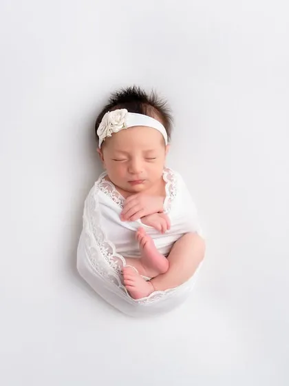 A newborn is swaddled in white lace, curled up against a pure white background. This high-key, minimalist photo emphasizes the baby's tiny form and the intricate details of the wrap.