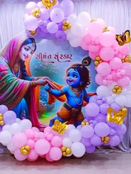 A traditional and divine baby shower theme featuring Lord Krishna. The circular backdrop with beautiful artwork is surrounded by a garland of pink, purple, and white balloons.
