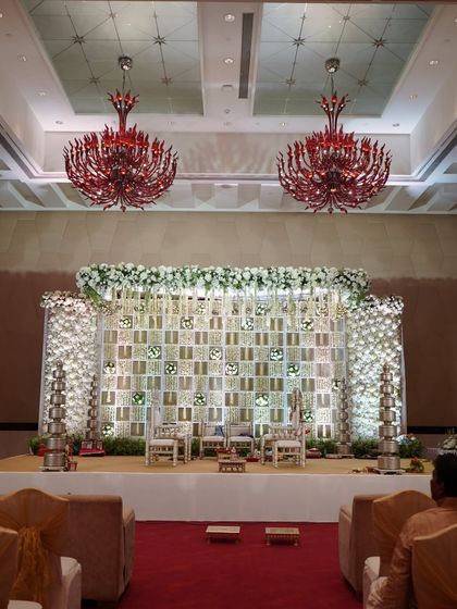This elegant stage features a backdrop of white jaali panels with niches for traditional tiered lamps (diyas). The all-white theme creates a serene and sophisticated atmosphere for a wedding ceremony or reception.