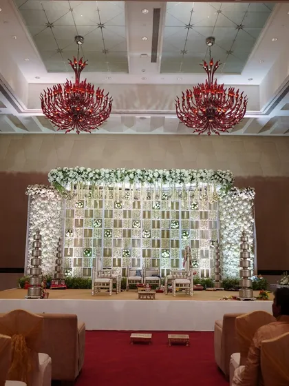 A stunning wedding stage featuring a backdrop of intricate white lattice panels, traditional silver 'diyas', and red chandeliers, blending modern and classic elements.