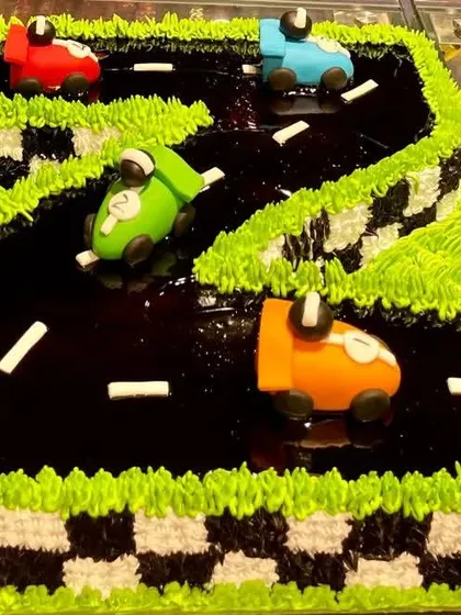 A creative race car track cake sculpted into the shape of the number two. This design features a chocolate ganache track, buttercream grass, and colorful fondant cars, perfect for a second birthday.