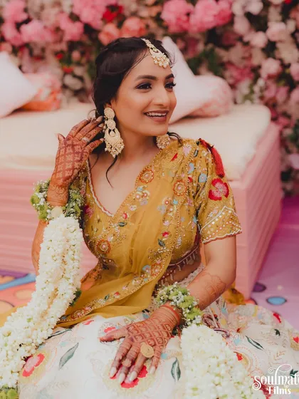A beautiful portrait of a bride at her Haldi ceremony. She is adorned with floral jewelry, sitting against a backdrop of flowers, radiating happiness.