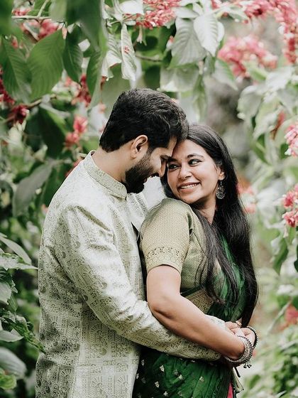 A romantic embrace amidst a garden of pink flowers, the couple dressed in beautiful green and cream traditional wear.