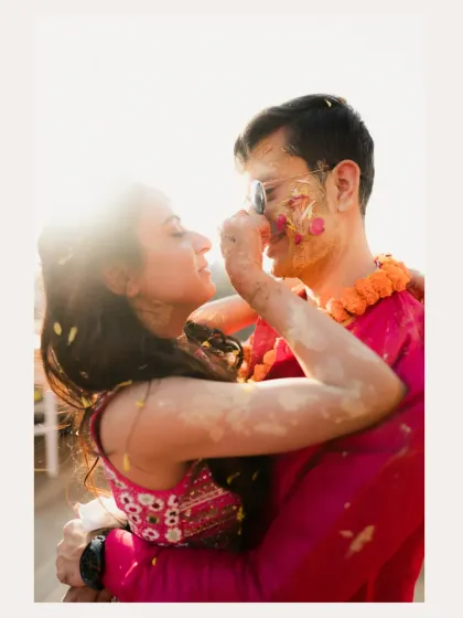 A tender moment where the bride playfully applies Haldi to the groom's face, backlit by the sun.