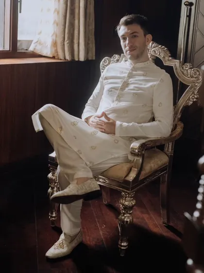 A relaxed portrait of the groom seated in an ornate, gilded chair, looking confidently at the camera in his elegant white sherwani.