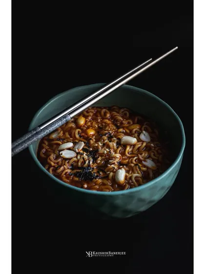 A bowl of spicy ramen noodles, shot with a minimalist, dark photography style. The focus is entirely on the food, using a single light source to highlight the texture of the noodles and toppings.