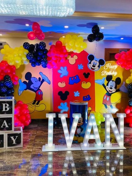 A vibrant and grand Mickey Mouse themed first birthday. This large-scale setup includes multiple character backdrops, colorful balloon arrangements, light-up letters for the name 'IVAN', and a marquee number '1'.