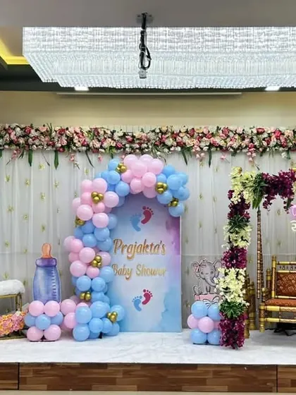 A combined baby shower and naming ceremony stage. It features a floral moon seat, a decorated jhula, and a pink and blue balloon arch.
