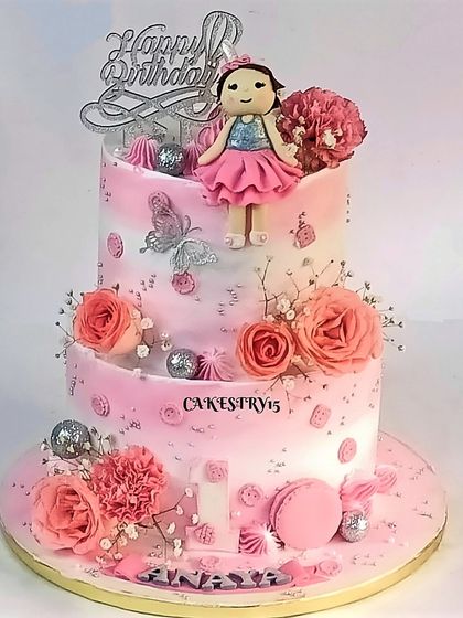 A lovely two-tier pink cake for a first birthday. It features a fondant doll topper, real and sugar flowers, macarons, and silver butterfly accents for an elegant and sweet design.