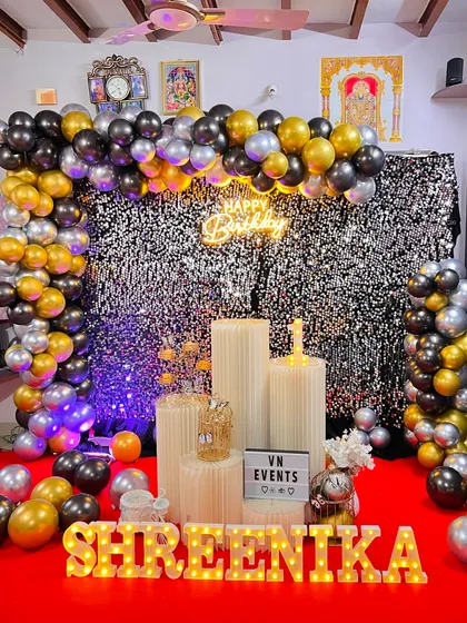 A different lighting angle on the black, silver, and gold birthday setup against a shimmer wall. The blue uplighting adds a cool contrast to the warm gold balloons.