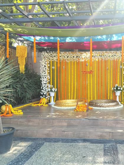 An outdoor Haldi setup on a wooden deck. The backdrop features yellow and orange drapes with strings of white and yellow flowers, and two brass urulis for the ceremony.