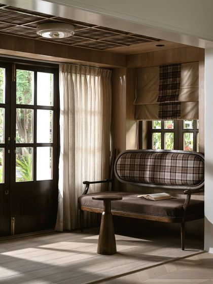 Adjacent to the office, a reading nook is furnished with a custom bench upholstered in plaid fabric, paired with a small, sculptural side table. The woven wood ceiling adds another layer of craftsmanship.