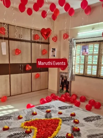 A romantic bedroom setup with red balloons on the ceiling and floor, and a heart made of flower petals on the bed.