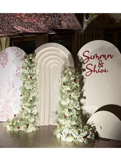 A modern and chic backdrop for an anniversary celebration. This setup features contemporary arched panels, one with a custom floral print and another with the couple's names, adorned with beautiful white floral arrangements.
