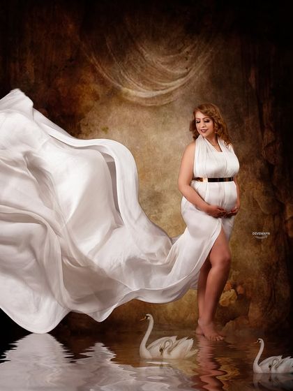 A whimsical and graceful portrait featuring a flowing white gown, swan props, and a water reflection. The fabric toss creates a beautiful sense of motion, like a gentle wave.