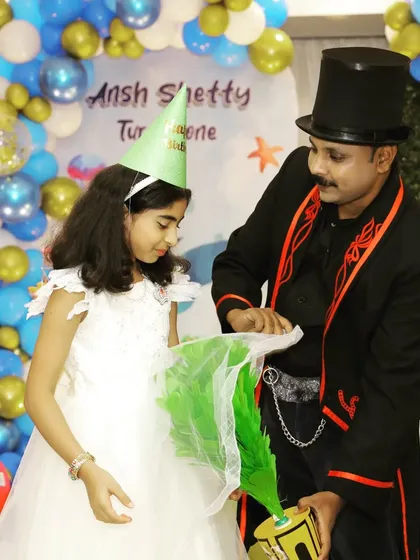 The birthday girl assists me with a trick involving colorful props at Ansh Shetty's first birthday party. I always invite the kids to participate in the magic.