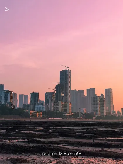 A 2x zoom shot of the Mumbai skyline, offering a balanced composition that bridges the gap between a wide view and a close-up. This mobile photo shows the city's profile against a soft, hazy sunset.