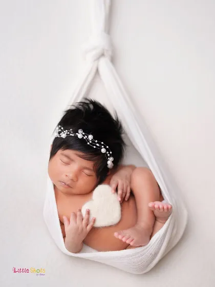 A creative and artistic shot of a baby sleeping in a white fabric sling, holding a small heart. This pose is beautiful, simple, and elegant.