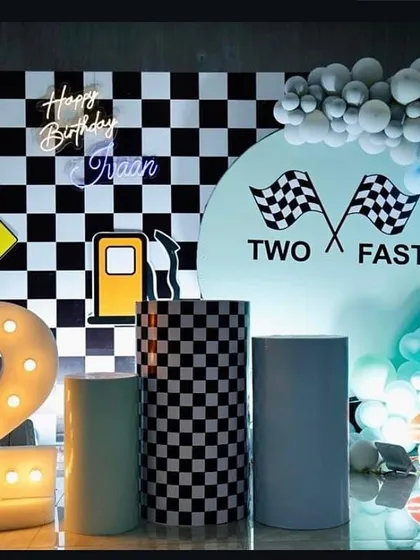 A "Two Fast" car-themed second birthday party. The setup includes a checkered flag backdrop, a custom "Two Fast" sign, a light-up number '2', and a blue balloon garland to complete the race-day look.