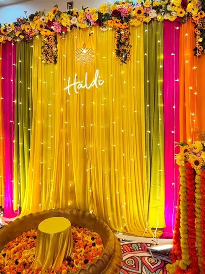 A vibrant Haldi setup featuring a large urli for the ceremony. The backdrop is a mix of colorful drapes, a "Haldi" neon sign, and fairy lights, creating a bright and festive atmosphere.