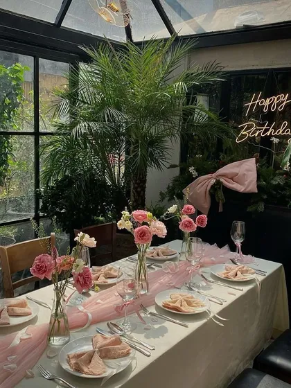 This pretty-in-pink birthday dinner was set up in a beautiful glass conservatory. The decor features a soft pink table runner, matching napkins, and fresh pink roses, all illuminated by a custom "Happy Birthday" neon sign for a personal touch.