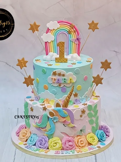 A magical two-tier unicorn-themed cake for a first birthday. It features a fondant unicorn head, a rainbow, and colorful roses, all in a soft pastel palette.