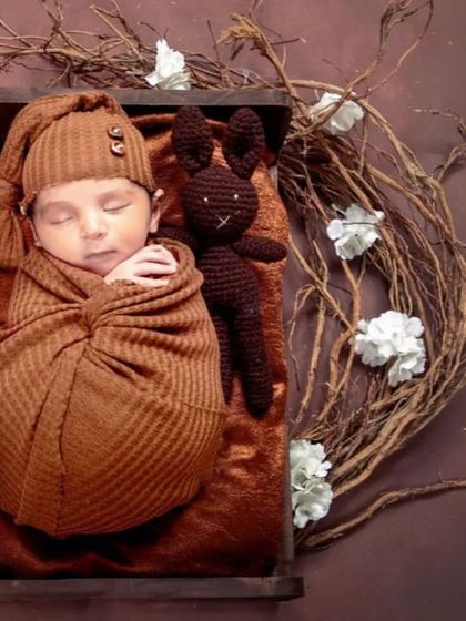 A rustic and warm newborn setup with a spring wreath theme. The baby is swaddled in brown and placed in a small wooden bed, accompanied by a knitted bunny toy and surrounded by natural twigs.