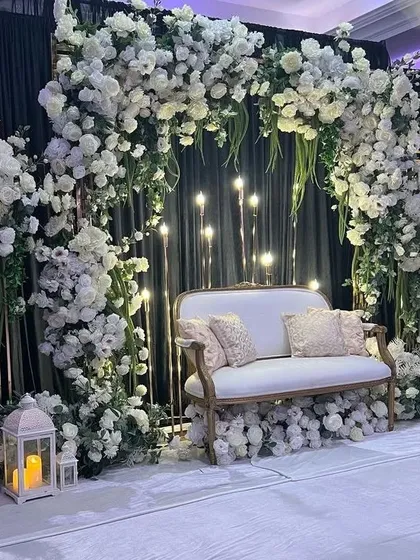 A luxurious black and white themed stage. The decor features a black drape backdrop, a profusion of white flowers creating an arch, and a vintage white sofa, all illuminated by candles in lanterns.