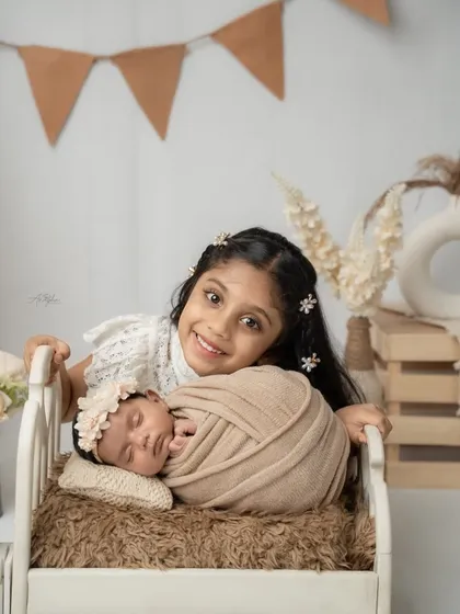 A heartwarming moment between siblings, where an older sister lovingly smiles over her newborn sibling sleeping in a tiny rustic bed.