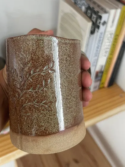 A close-up of a hand-built mug with a subtle, embossed leaf pattern. The speckled glaze gives it a rustic, earthy quality.